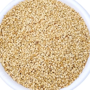 May include: A close-up shot of a white bowl filled with sesame seeds. The seeds are small, oval-shaped, and beige in color. The bowl is white and the seeds fill the entire frame, creating a textured surface.
