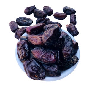 Pode incluir: Uma tigela branca cheia de tâmaras marrons escuras. As tâmaras têm uma textura enrugada e variam em tamanho e forma. Algumas tâmaras são de uma cor marrom avermelhada escura. A tigela está em um fundo branco.
