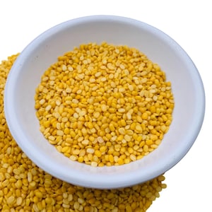 Puede incluir: Un cuenco blanco lleno de frijoles mungo amarillos partidos, con más frijoles derramándose alrededor del cuenco. Los frijoles son de un color amarillo vibrante, y el cuenco tiene un diseño simple y limpio.