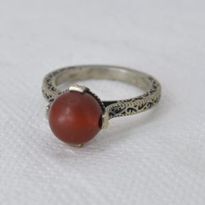 Può includere: Un anello d'argento con una fascia ornata e una grande pietra rotonda marrone-rossastra. La fascia dell'anello presenta un motivo a spirale dettagliato. La pietra è incastonata in un'incastonatura quadrata semplice.
