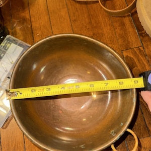 May include: A round copper bowl with a smooth interior and a metallic finish. A yellow measuring tape is stretched across the diameter, indicating a width of approximately 9 inches. The bowl features a small circular handle on the side.