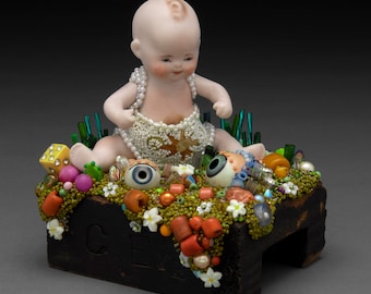 May include: A porcelain baby doll sits on a wooden block covered in colorful beads, sequins, and other embellishments. The doll is wearing a white dress with a beaded design and has a star on its chest. The block has the word "CELL" written on it.