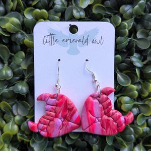 May include: A pair of handmade earrings featuring a witch hat design. The earrings are made of marbled polymer clay in shades of pink and red, with silver-colored ear wires. The card behind the earrings reads "little emerald owl".