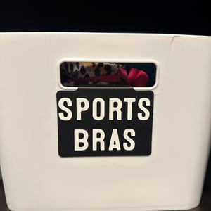 May include: A white storage bin with a black label that reads "SPORTS BRAS" in white capital letters. The bin has a rectangular opening, revealing a glimpse of sports bras in various patterns and colors.