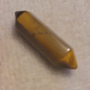 Może przedstawiać: Polerowany kryształ tygrysiego oka z dwoma zakończeniami. Kamień szlachetny ma złoto-brązowy kolor z błyszczącym wykończeniem. Kryształ ma kształt sześciokątnego pryzmatu, z ostrymi końcami.