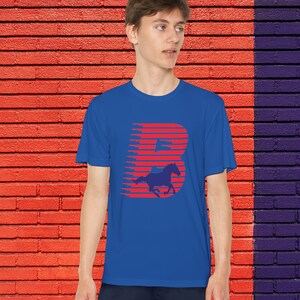 May include: A royal blue t-shirt with a red graphic of the letter "B" and a horse silhouette. The design uses horizontal lines for a dynamic look. The shirt is worn by a person, set against a brick wall with red and purple sections.