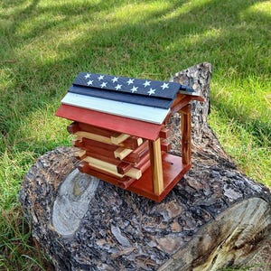 May include: A wooden birdhouse with a patriotic design. The roof is painted with the American flag, featuring blue with white stars, white, and red stripes. The birdhouse is made of stacked wooden logs and sits on a wooden base.