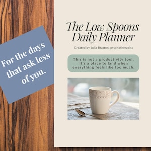 Può includere: Un'agenda giornaliera color crema intitolata "The Low Spoons Daily Planner" con il testo "For the days that ask less of you." L'agenda include una foto di una tazza bianca e un cucchiaio. L'agenda è su una superficie di legno.
