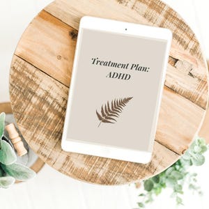 May include: A white tablet shows the text "Treatment Plan: ADHD" and a fern illustration. The tablet is on a round, light brown wooden surface, with greenery around it.