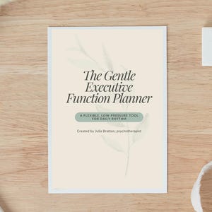 May include: A planner titled "The Gentle Executive Function Planner" with text that reads "A flexible, low-pressure tool for daily rhythm." The planner is on a light wood surface.