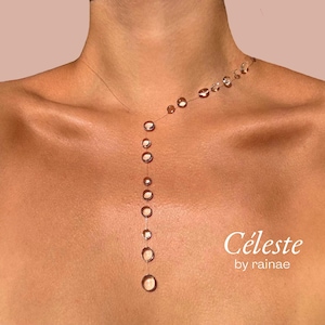 Peut inclure: Un collier délicat composé d'un cordon fin et transparent orné d'une série de petites perles circulaires et claires qui descendent en ligne verticale depuis la clavicule. Le texte Céleste by rainae est imprimé en blanc en bas à droite.