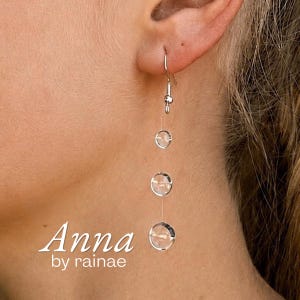 May include: A silver-toned drop earring featuring three circular, clear, faceted stones suspended vertically on a thin wire. The earring is attached to a silver hook. The text "Anna by rainae" is printed in white in the lower left corner.