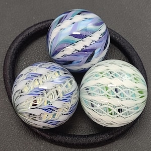May include: Three spherical glass marbles featuring intricate swirling patterns of white, blue, and light green glass filaments. The marbles are arranged in a cluster, resting on a circular black elastic hair tie, showcasing smooth, reflective surfaces.