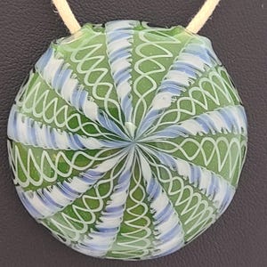 May include: A circular glass pendant featuring a vibrant green base with intricate white and light blue swirling patterns that radiate from the center. The design includes delicate, interconnected loops and lines, suspended by a light-colored cord.