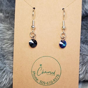 May include: A pair of dangle earrings with dark blue, round crystal pendants. The earrings hang from silver-colored hooks and are displayed on a brown card with the "Charmed Jewelry" logo and phone number.