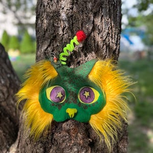 May include: A whimsical, handmade plush alien face with yellow fur, green skin, and purple star-shaped eyes. It has a green and red antenna and is set against a tree trunk.
