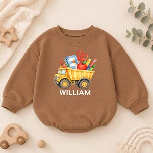 May include: A brown long-sleeve sweatshirt featuring a graphic of a yellow dump truck carrying school supplies including a blue notebook, red scissors, a red apple, glue, and colorful pencils, with the name WILLIAM printed in white below the truck.
