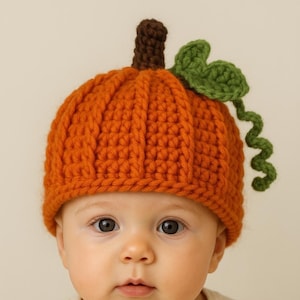 May include: A baby wearing an orange crocheted pumpkin hat with a brown stem and green leaf accents. The hat is designed to resemble a pumpkin. The baby is wearing a white onesie.