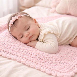 Könnte beinhalten: Ein schlafendes Baby mit einem floralen Stirnband und einem cremefarbenen Strampler, das auf einer hellrosa gehäkelten Decke ruht. Die Decke hat einen gewellten Rand. Das Baby liegt auf einem Bett mit weißem Laken und rosa Kissen.