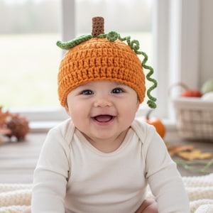 May include: A smiling baby wearing an orange crochet pumpkin hat with green vines and a brown stem. The baby is wearing a white long-sleeved onesie and sitting on a cream-colored blanket.