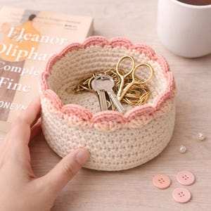May include: A cream-colored, crocheted basket with a scalloped pink trim. The basket contains keys and gold scissors. A book titled "Eleanor Oliphant is Completely Fine" is in the background, along with a cup of tea and pink buttons.