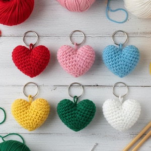 May include: Six handmade crochet heart keyrings in red, pink, blue, yellow, green, and white. Each heart is attached to a silver keyring. Yarn balls in matching colours are arranged around the keyrings on a white wooden surface.