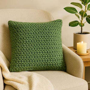 May include: A square, green crochet pillow rests on a beige armchair. The pillow has a textured surface and a solid green color. The chair is next to a small wooden table with a lit candle and a potted plant.
