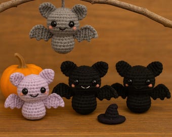 May include: A collection of handmade crochet bats in various colors, including gray, black, and lavender. One bat hangs from a branch, while others sit on a wooden surface. A small pumpkin and a witch hat are also present, creating a Halloween theme.