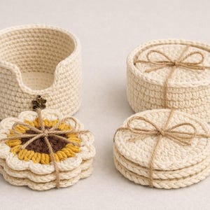 Könnte beinhalten: Eine Sammlung handgefertigter Häkelartikel in Creme und Gelb. Das Set beinhaltet einen Halter, zwei runde Behälter und zwei Stapel Untersetzer. Die Untersetzer sind sonnenblumenförmig und mit Bindfaden gebunden. Der Halter hat einen dekorativen Metallakzent.