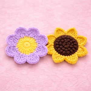 Può includere: Due fiori fatti a mano all'uncinetto. Uno è viola con un centro giallo, l'altro è un girasole giallo con un centro marrone. I fiori sono su uno sfondo rosa. Questi fiori decorativi sono perfetti per l'artigianato.