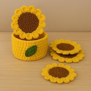 Puede incluir: Un juego de posavasos de girasol de ganchillo hechos a mano y un recipiente a juego. Los posavasos y el recipiente son amarillos con centros marrones, y el recipiente tiene un acento de hoja verde. Los posavasos tienen unos 10 cm de diámetro.