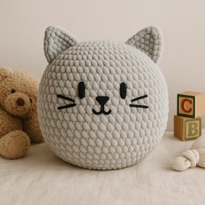 Puede incluir: Una almohada redonda gris claro con forma de gato con rasgos faciales bordados en negro, incluyendo ojos, nariz y bigotes. La almohada tiene dos pequeñas orejas triangulares. Un oso de peluche y bloques de madera están en el fondo.