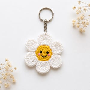 May include: A handmade crochet daisy keychain with a smiling face. The daisy has white petals and a yellow center with pink cheeks and a black smile. The keychain is attached to a silver metal ring.