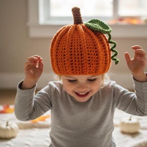 Peut inclure: Un chapeau citrouille en crochet orange, avec une tige marron et une feuille verte. Le chapeau est porté par un enfant vêtu d'un haut gris à manches longues. Un accessoire de saison pour l'automne ou Halloween.