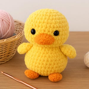 Op de afbeelding: Een handgemaakte gele gehaakte eend met zwarte ogen en een oranje snavel en voeten. Een mand met roze en blauwe garenballen staat achter de eend. Een haaknaald en een bolletje crèmekleurig garen zijn ook aanwezig.