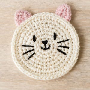 May include: A round, off-white crochet coaster with a cat face design. The cat face features black embroidered eyes, nose, and whiskers, with two small pink ears. The coaster is made of a soft, textured yarn.