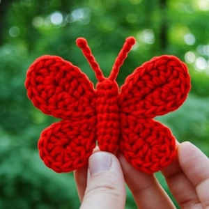 May include: A vibrant red crocheted butterfly with detailed wings and antennae. The handmade craft is held against a blurred green background, showcasing the intricate stitch work and the butterfly's symmetrical design. A charming decorative item.