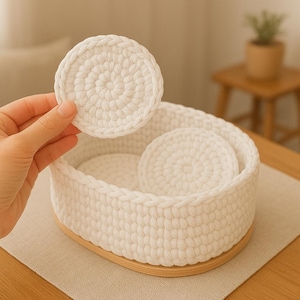 Puede incluir: Una cesta blanca de ganchillo con base de madera, que contiene varias almohadillas redondas blancas de ganchillo. Una almohadilla está siendo sostenida por una mano. La cesta y las almohadillas están hechas de un hilo grueso y texturizado.
