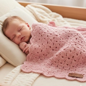 Op de afbeelding: Een slapende baby ligt op een kussen, bedekt met een roze gehaakte deken. De deken heeft een geschulpte rand en een textuurpatroon. Een klein houten label op de deken leest "Handmade With Love".