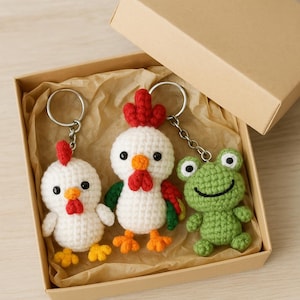 Puede incluir: Una caja de regalo con tres llaveros de ganchillo. Los llaveros tienen forma de un pollo blanco con detalles rojos y naranjas, y una rana verde. La caja es de cartón y contiene papel marrón.