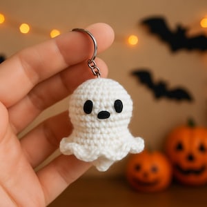 Peut inclure: Un porte-clés fantôme blanc crocheté à la main, avec des yeux et une bouche noirs. Le fantôme a une tête ronde et un corps fluide. Le porte-clés est attaché à un anneau en métal argenté. L'arrière-plan comprend des décorations d'Halloween.