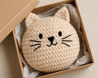 May include: A beige crocheted cat-shaped pillow with black embroidered facial features, including eyes, nose, and whiskers, is presented inside a brown cardboard box. The pillow is round and has two pointed ears. The box is open, revealing the pillow nestled on white tissue paper.