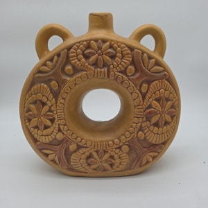 Könnte beinhalten: Eine beige Keramikflasche mit rundem Körper und einem zentralen Loch. Die Flasche weist aufwendige florale und geometrische Reliefmuster, zwei seitliche Griffe und einen kleinen Ausguss auf. Das Design ist symmetrisch und dekorativ.