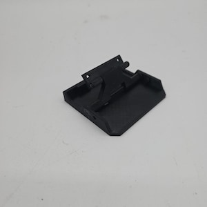 May include: A black 3D-printed object with a rectangular base and an angled top section. The object has a hinged mechanism and several small holes. The overall design is geometric and functional.