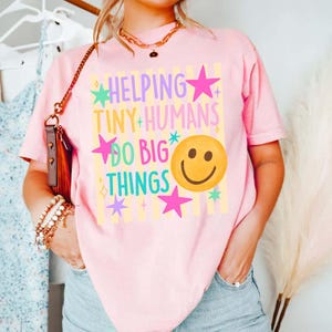 Può includere: Una t-shirt rosa chiaro con una faccina sorridente gialla e il testo "HELPING TINY HUMANS DO BIG THINGS" in lettere colorate. Il design include strisce verticali gialle e varie stelle multicolori e piccoli accenti a forma di croce.