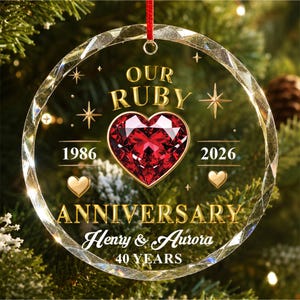 May include: A round, faceted glass ornament featuring a central red heart graphic. Text reads: OUR RUBY, 1986, 2026, ANNIVERSARY, Henry & Aurora, 40 YEARS. The design includes gold lettering, small star accents, and two small gold hearts, suspended by a red ribbon.