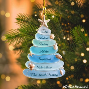 May include: A flat, tree-shaped Christmas ornament featuring stacked, smooth, blue-toned glass pieces. The ornament is topped with a starfish and decorated with small seashells and pearls. It displays the text 2026, Aiden, Daisy, Adela, Nymeria, Christian, and The Smith Family.