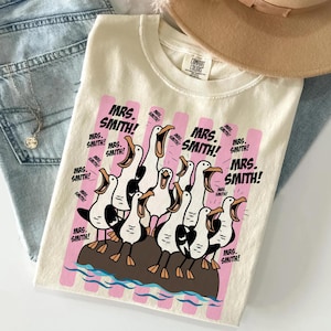 May include: A cream-colored t-shirt featuring a graphic of multiple seagulls standing on a brown rock with blue water waves below. The text "MRS. SMITH!" is repeated multiple times in black font over vertical pink stripes on the shirt front.