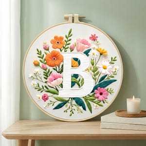 以下が含まれることがあります： カラフルな花に囲まれた大きな白い文字「B」が特徴の刺繍フラワーフープアート。 花には、オレンジ、ピンク、白の花と緑の葉が含まれています。 フープは木製の表面に表示されています。