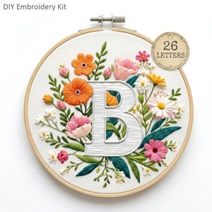 May include: Embroidery kit featuring a floral design with the letter "B" in white, surrounded by orange, pink, and white flowers, green leaves, and the text "26 LETTERS" on a tag. The embroidery is set in a wooden hoop.